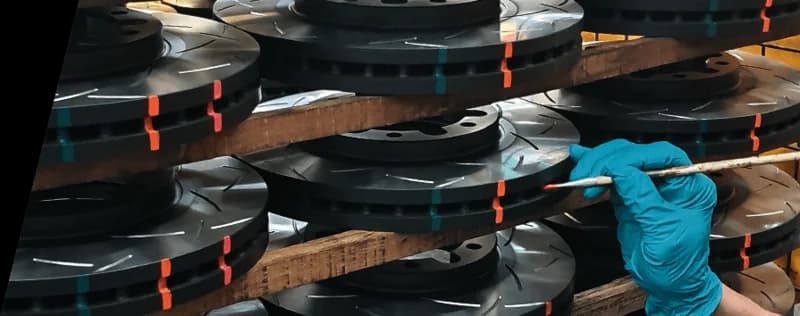 Technician in blue gloves inspecting stacked DBA slotted brake rotors with colorful temperature indicator markings during quality control process