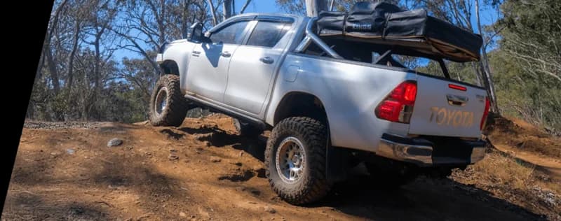 White Toyota pickup truck loaded with camping gear positioned on rocky terrain in Australian bush demonstrating heavy load off-road conditions